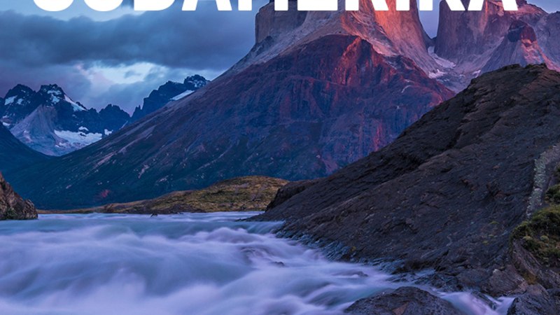 Ein Fluss fließt durch eine felsige Landschaft, im Hintergrund erheben sich majestätische Berge unter einem dramatischen Himmel. Text: SÜDAMERIKA., © Expedition Erde