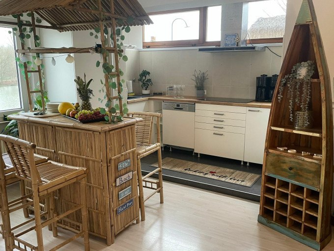 Helle Küche mit Bambusbar, dekoriert mit Früchten und Pflanzen. Ein Regal in Bootsform steht rechts, Fenster mit Blick auf Wasser im Hintergrund., © Cool-Tours StattReisen Helle Küche mit Bambusbar, dekoriert mit Früchten und Pflanzen. Ein Regal in Bootsform steht rechts, Fenster mit Blick auf Wasser im Hintergrund., © Cool-Tours StattReisen