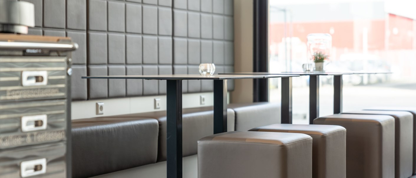 Moderner Fr&uuml;hst&uuml;cksbereich mit gepolsterten Sitzb&auml;nken, W&uuml;rfelhockern und einem Tisch. Im Hintergrund ist ein Fenster mit Blick nach drau&szlig;en., &copy; a2 HOTELS