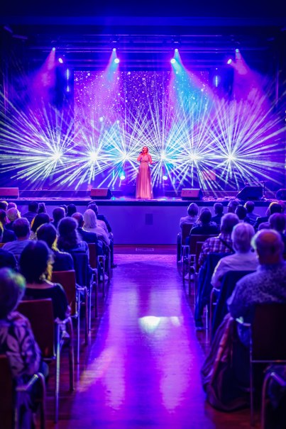 Eine Sängerin steht auf der Bühne der Stadthalle K3N, umgeben von bunten Lichteffekten. Das Publikum sitzt in Reihen und schaut zu., © Daniel Jueptner | 8pm.de