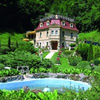 Ein kleines Schloss mit rotem Dach, umgeben von einem gepflegten Garten mit Springbrunnen und üppiger Vegetation.
