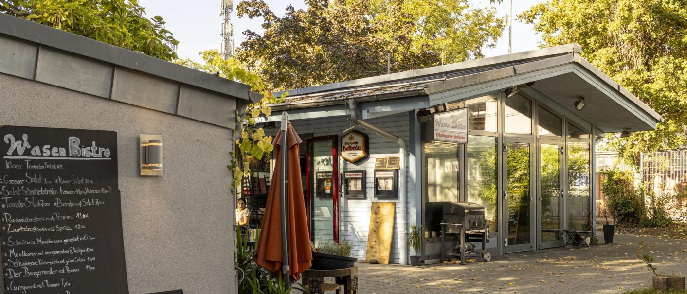 Ein kleines Bistro mit Glasfront und Menütafel. Ein Sonnenschirm steht davor, umgeben von Bäumen und Pflanzen., © Stuttgart-Marketing GmbH, Sarah Schmid