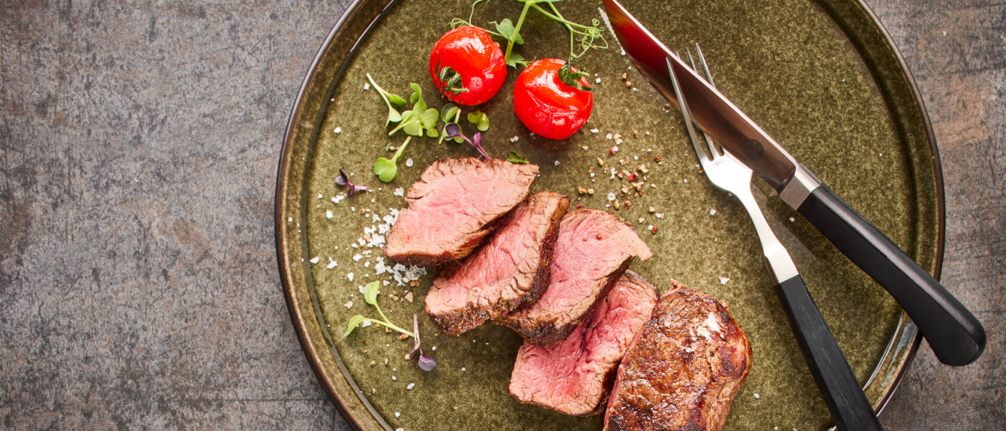 Ein Teller mit geschnittenem Steak, zwei Kirschtomaten und Besteck auf einem rustikalen Untergrund., © MAREDO