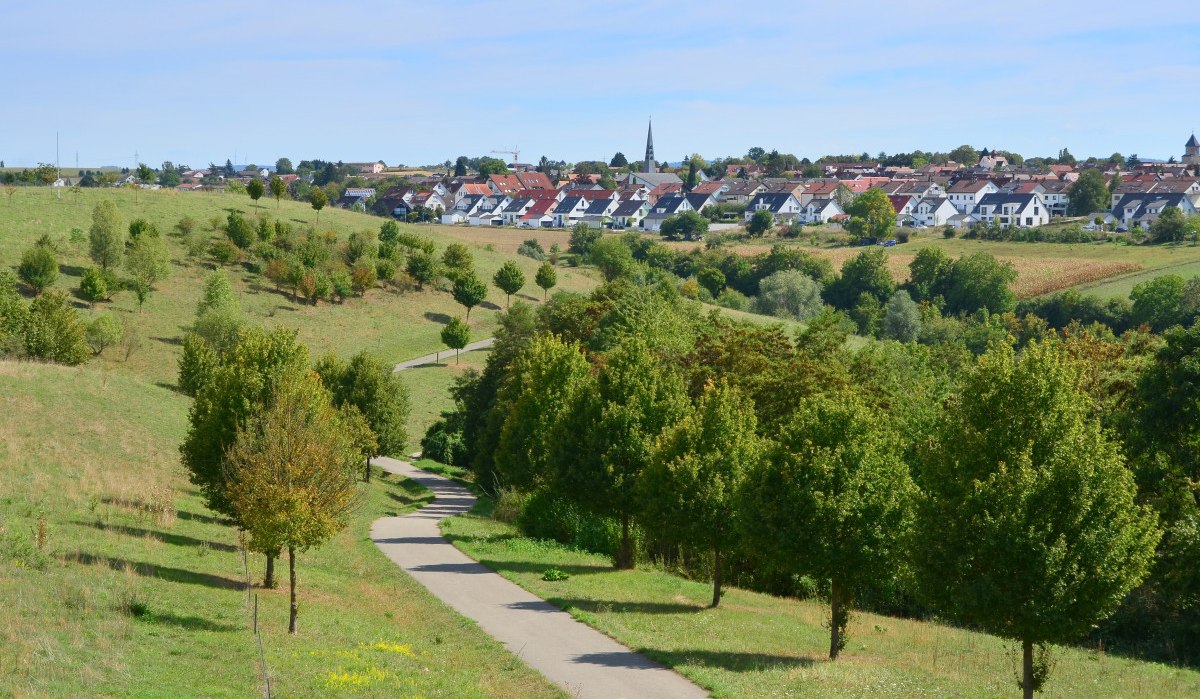 Ein gewundener Weg führt durch eine grüne Landschaft mit Bäumen. Im Hintergrund ist eine Siedlung mit Häusern und einer Kirche sichtbar., © Remstal Tourismus e.V. Ein gewundener Weg führt durch eine grüne Landschaft mit Bäumen. Im Hintergrund ist eine Siedlung mit Häusern und einer Kirche sichtbar., © Remstal Tourismus e.V.