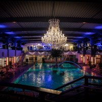 Luxuriöses Spa mit großem Kronleuchter über einem beleuchteten Pool, umgeben von Liegestühlen und Pflanzen. Elegante Atmosphäre mit blauer Beleuchtung., © AlbThermen Bad Urach