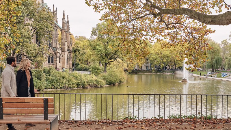 Ein Paar spaziert am Feuersee entlang, im Hintergrund die Johanneskirche und herbstliche B&auml;ume. Ein Brunnen sprudelt im See., &copy; SMG Christoph D&uuml;pper