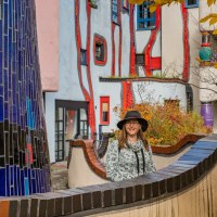 Frau mit Hut vor dem farbenfrohen Hundertwasserhaus in Plochingen, umgeben von herbstlichen Blättern., © Bildergalerie Attila