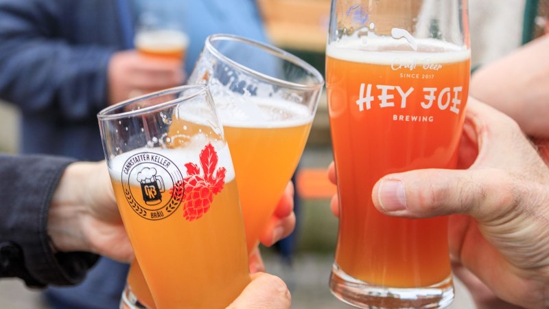Drei Personen stoßen mit Biergläsern an, die Logos von "Hey Joe Brewing" und "Cannstatter Keller Bräu" tragen. Drei Personen stoßen mit Biergläsern an, die Logos von "Hey Joe Brewing" und "Cannstatter Keller Bräu" tragen.
