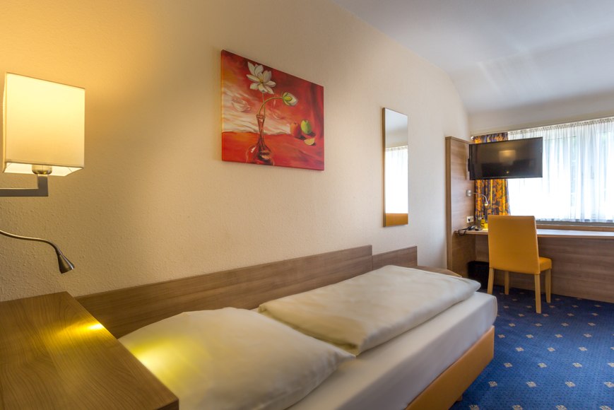 Ein gemütliches Einzelzimmer mit einem Bett, einem Schreibtisch mit gelbem Stuhl, einem Fernseher und einem Fenster mit Vorhängen., © Römerhof Hotelbetriebs GmbH