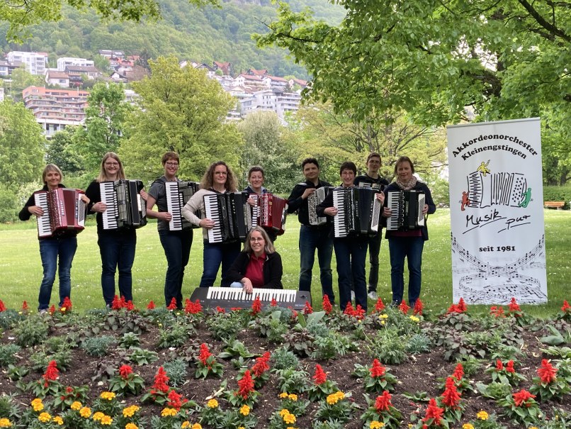 Das Akkordeonorchester Kleinengstingen posiert mit Akkordeons in einem blühenden Park. Ein Banner mit dem Orchesterlogo ist sichtbar., © AKK Kleinengstingen
