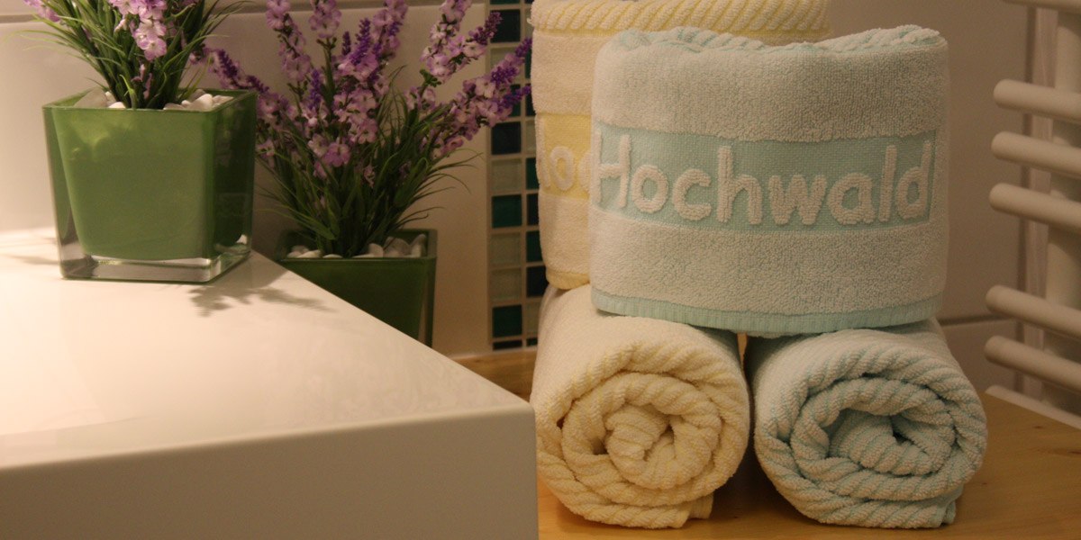 Badezimmer mit gefalteten Handtüchern, eines mit 'Hochwald' bestickt, und zwei grünen Pflanzgefäßen mit lila Blumen., © Hotel Hochwald