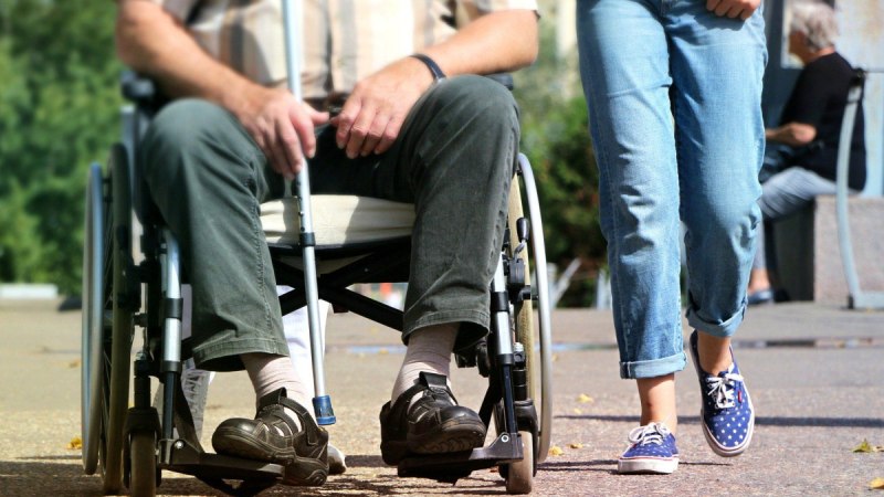 Ein Mann sitzt in einem Rollstuhl auf einem Gehweg, während eine Person in Jeans und Turnschuhen daneben geht., © pixabay