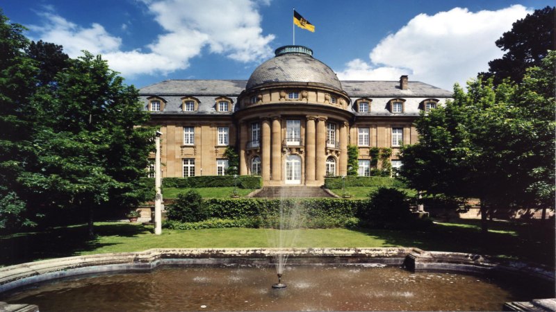 Die Villa Reitzenstein mit imposanter Fassade, einem Brunnen im Vordergrund und einer Flagge auf dem Dach, umgeben von Bäumen., © Staatsministerium Baden-Württemberg