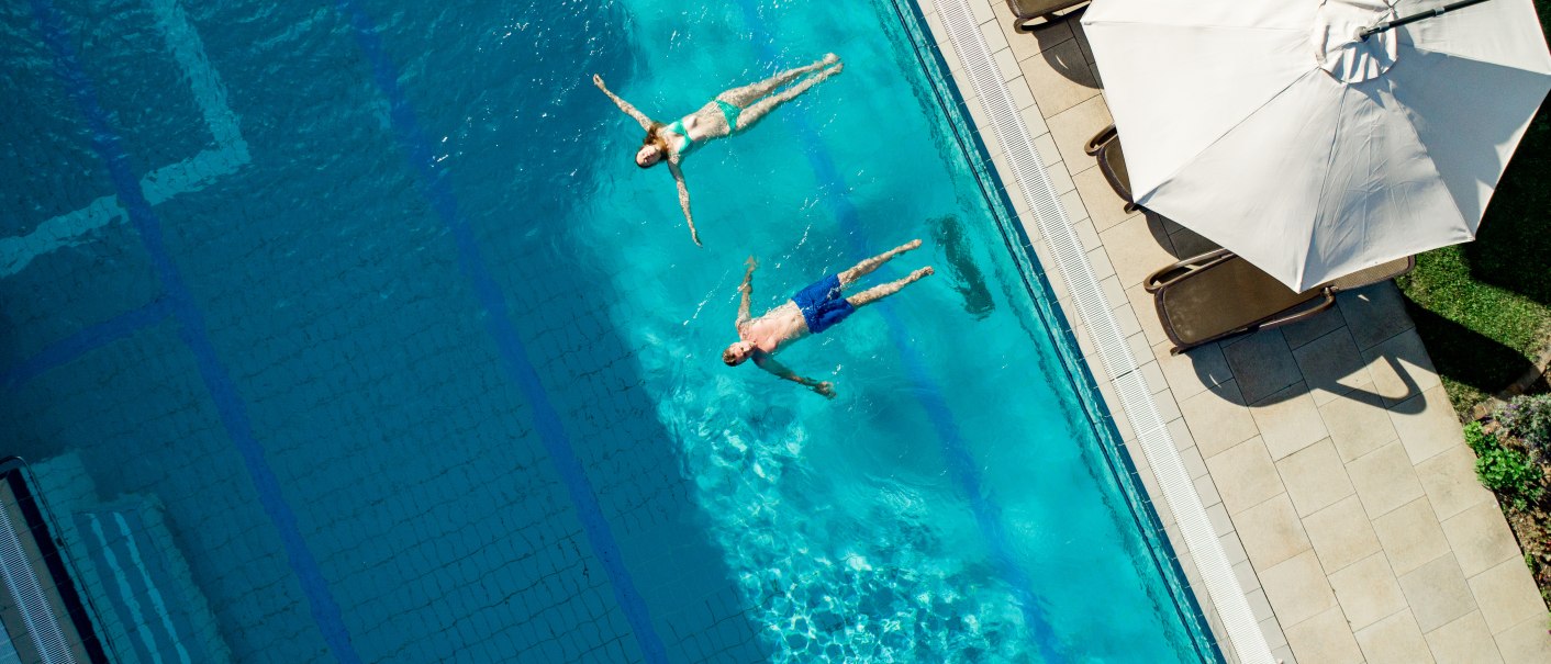 Zwei Personen treiben entspannt im blauen Wasser eines Pools. Daneben sind Liegestühle und ein großer Sonnenschirm zu sehen., © Christine Biedermann & Wolfgang Heiler
