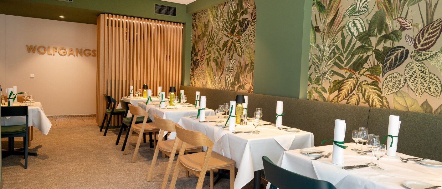 Restaurant mit grüner Wand und Pflanzenmotiv, gedeckte Tische mit weißen Tischdecken und Holzstühlen, Schriftzug 'WOLFGANGS' an der Wand., © Erikson Hotel Restaurant mit grüner Wand und Pflanzenmotiv, gedeckte Tische mit weißen Tischdecken und Holzstühlen, Schriftzug 'WOLFGANGS' an der Wand., © Erikson Hotel