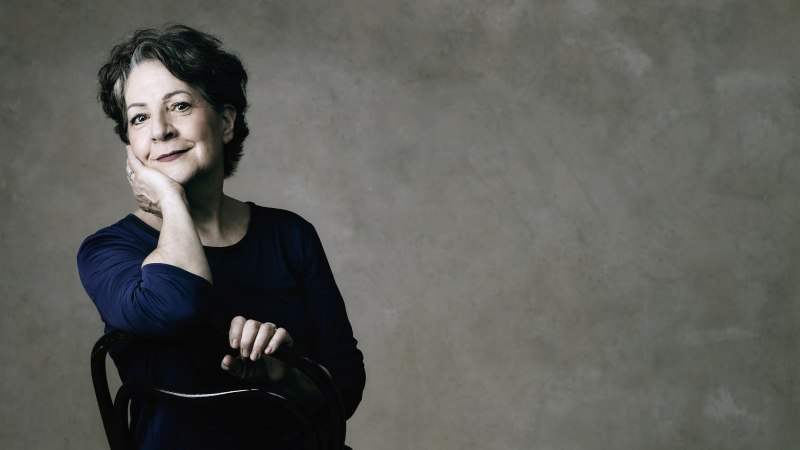 Frau in dunkler Kleidung sitzt lächelnd auf einem Stuhl vor grauer Wand.