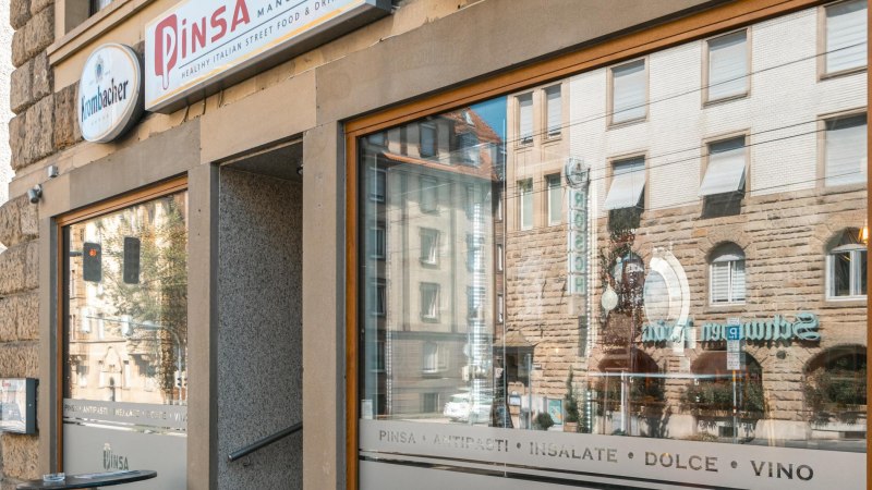 Außenansicht eines Restaurants mit der Aufschrift 'Pinsa Manufaktur' und Spiegelung von Gebäuden im Fenster., © SMG, Sarah Schmid