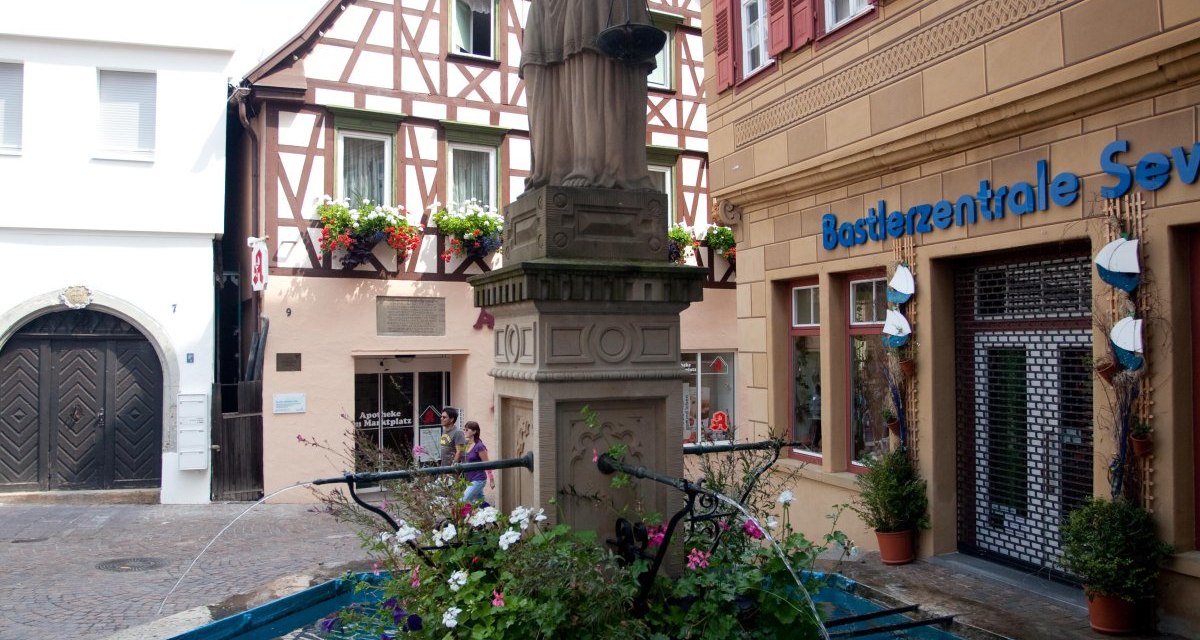 Justitia-Statue auf einem Marktbrunnen, umgeben von Fachwerkhäusern und einem Geschäft. Der Brunnen ist mit Blumen geschmückt., © Waiblingen - Stuttgart-Marketing GmbH Justitia-Statue auf einem Marktbrunnen, umgeben von Fachwerkhäusern und einem Geschäft. Der Brunnen ist mit Blumen geschmückt., © Waiblingen - Stuttgart-Marketing GmbH
