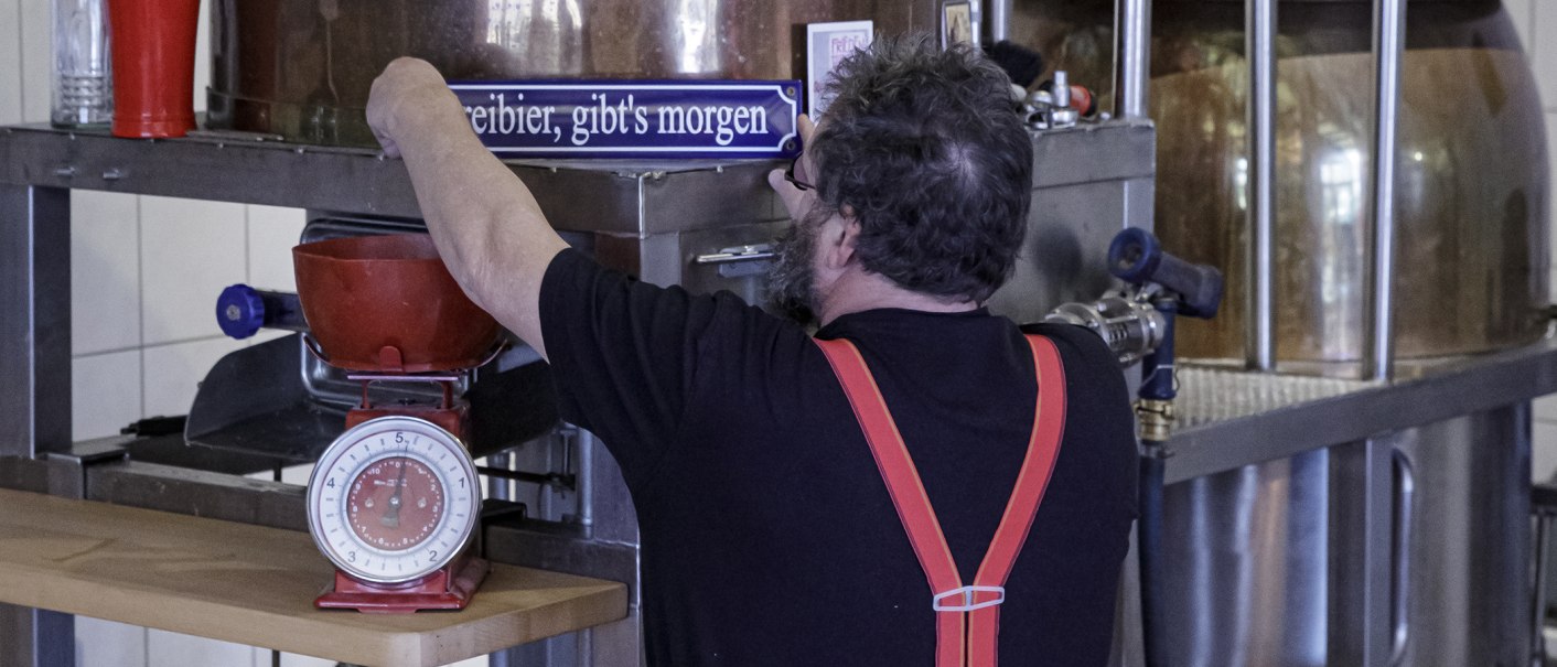 Ein Mann mit roten Hosenträgern arbeitet an einer Brauanlage in einer Gasthausbrauerei. Eine Waage und ein Schild sind sichtbar., © Torsten Wenzler Ein Mann mit roten Hosenträgern arbeitet an einer Brauanlage in einer Gasthausbrauerei. Eine Waage und ein Schild sind sichtbar., © Torsten Wenzler