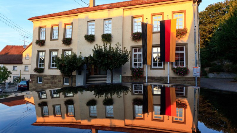 Das Rathaus in Waiblingen-Hohenacker, geschmückt mit Blumen und deutschen Flaggen, spiegelt sich auf einer glänzenden Oberfläche wider., © Waiblingen - Stuttgart-Marketing GmbH