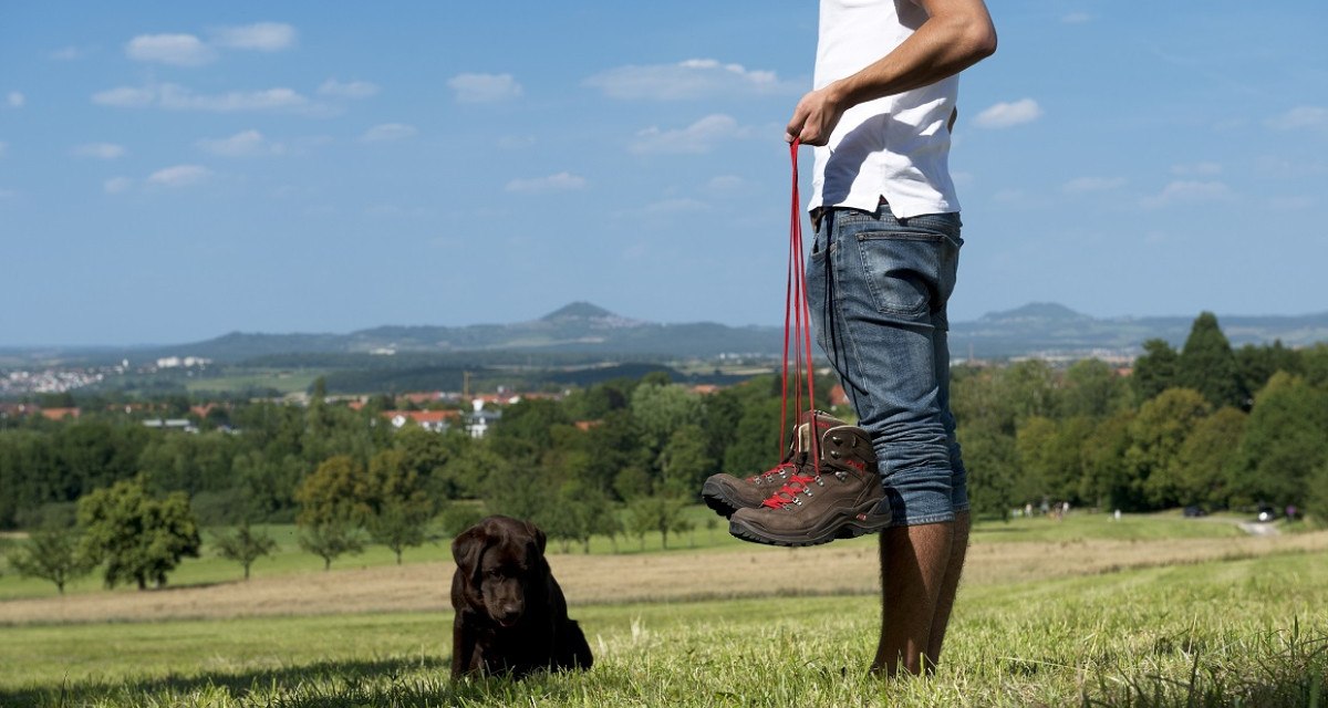 Person in Jeans und weißem T-Shirt hält Wanderschuhe. Ein Hund sitzt im Gras. Im Hintergrund sind grüne Felder und Hügel zu sehen., © Landkreis Göppingen Person in Jeans und weißem T-Shirt hält Wanderschuhe. Ein Hund sitzt im Gras. Im Hintergrund sind grüne Felder und Hügel zu sehen., © Landkreis Göppingen