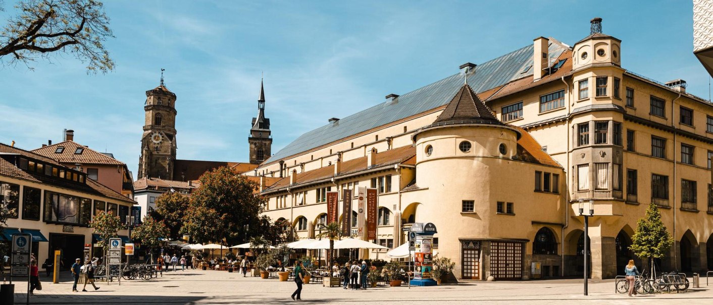 Die Markthalle Stuttgart bei sonnigem Wetter, umgeben von Menschen, Fahrrädern und Bäumen. Historische Gebäude im Hintergrund., © Stuttgart Marketing GmbH, Sarah Schmid