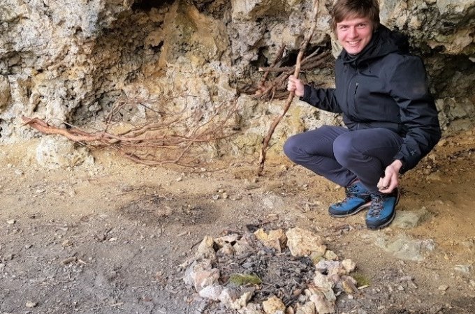 Eine Person in schwarzer Jacke kniet in einer Höhle vor einem Steinkreis. Sie hält einen Stock und lächelt in die Kamera., © SwabianTravel Eine Person in schwarzer Jacke kniet in einer Höhle vor einem Steinkreis. Sie hält einen Stock und lächelt in die Kamera., © SwabianTravel