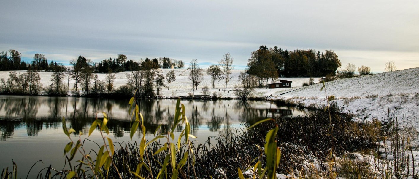 Winterliche Szene am Aichstruter Stausee mit schneebedeckten Feldern und Bäumen, die sich im ruhigen Wasser spiegeln. Ein kleines Gebäude steht am Ufer., © Stuttgart-Marketing GmbH, Sarah Schmid Winterliche Szene am Aichstruter Stausee mit schneebedeckten Feldern und Bäumen, die sich im ruhigen Wasser spiegeln. Ein kleines Gebäude steht am Ufer., © Stuttgart-Marketing GmbH, Sarah Schmid