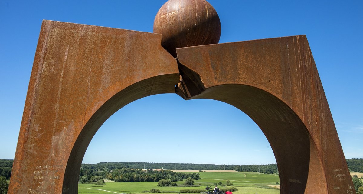 Rostige Metallskulptur mit Kugel auf einem Hügel, darunter grüne Landschaft und blauer Himmel. Ein Fahrradfahrer sitzt im Hintergrund., © Stuttgart Marketing GmbH Rostige Metallskulptur mit Kugel auf einem Hügel, darunter grüne Landschaft und blauer Himmel. Ein Fahrradfahrer sitzt im Hintergrund., © Stuttgart Marketing GmbH