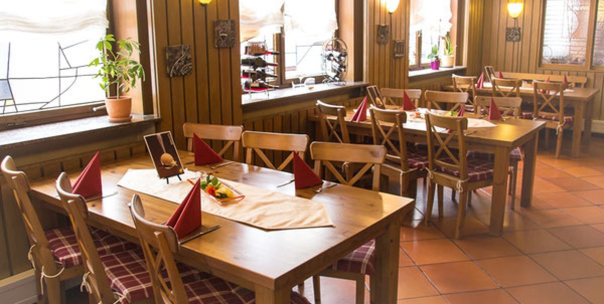 Gemütliche Gaststube mit Holztischen, Stühlen und roten Servietten. Die Fensterbank ist mit Pflanzen dekoriert, die Wände sind holzvertäfelt.