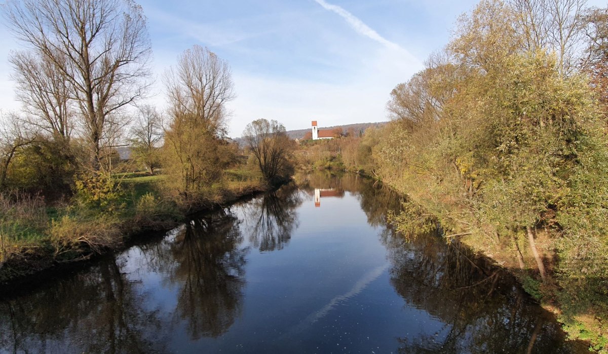 Ein ruhiger Fluss, gesäumt von kahlen Bäumen, spiegelt den blauen Himmel wider. Im Hintergrund ist eine Kirche mit rotem Dach zu sehen., © Kulturamt Plochingen