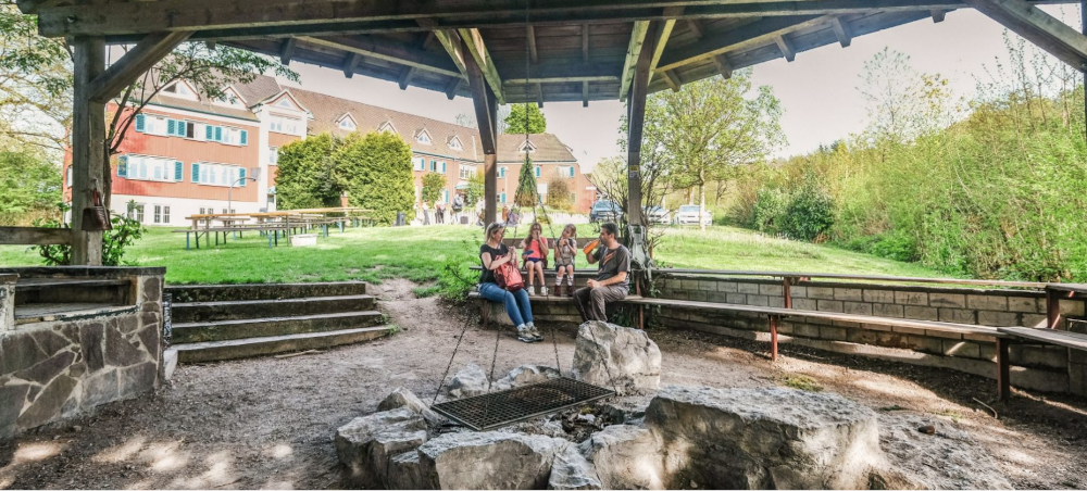 Überdachter Grillplatz mit Sitzbänken und Personen, im Hintergrund ein Gebäude mit blauen Fensterläden., © DJH Baden-Württemberg e.V.