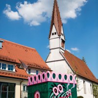 Bunt bemaltes Gebäude mit Drachenmotiv und pinken Details neben einer Kirche mit spitzem Turm in Plochingen., © Stuttgart-Marketing GmbH, Achim Mende Bunt bemaltes Gebäude mit Drachenmotiv und pinken Details neben einer Kirche mit spitzem Turm in Plochingen., © Stuttgart-Marketing GmbH, Achim Mende