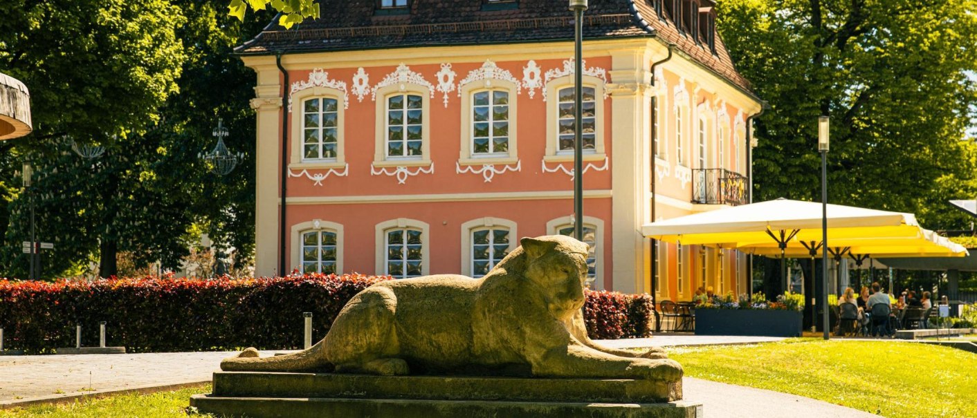 Das Rokokoschlösschen in Schwäbisch Gmünd, umgeben von Bäumen, mit einer Löwenstatue im Vordergrund und einem Cafébereich mit gelben Sonnenschirmen., © Stuttgart-Marketing GmbH, Sarah Schmid