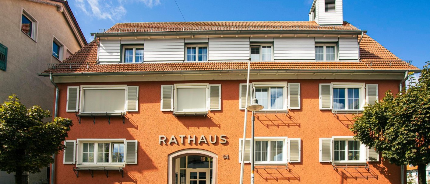 Rathaus von Bad Boll mit orangefarbener Fassade, weißen Fensterläden und kleinem Turm. Vor dem Gebäude stehen Bänke und Fahrräder, umgeben von Bäumen., © Stuttgart-Marketing GmbH, Sarah Schmid
