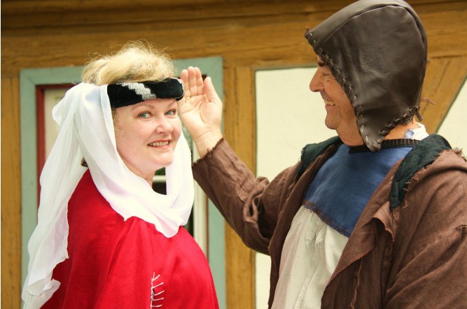 Zwei Personen in mittelalterlicher Kleidung lächeln vor einem Fachwerkhaus. Die Frau trägt ein rotes Gewand, der Mann eine braune Kapuze., © Touristik und Marketing GmbH