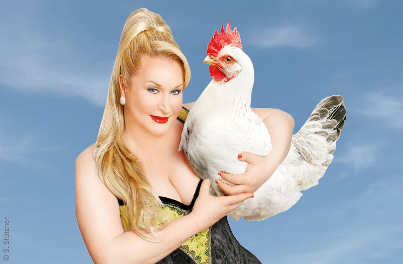 Eine Person mit blonden Haaren hält ein weißes Huhn vor einem blauen Himmel. Sie trägt ein elegantes Outfit und lächelt in die Kamera., © Stadt Kornwestheim Kulturmanagement