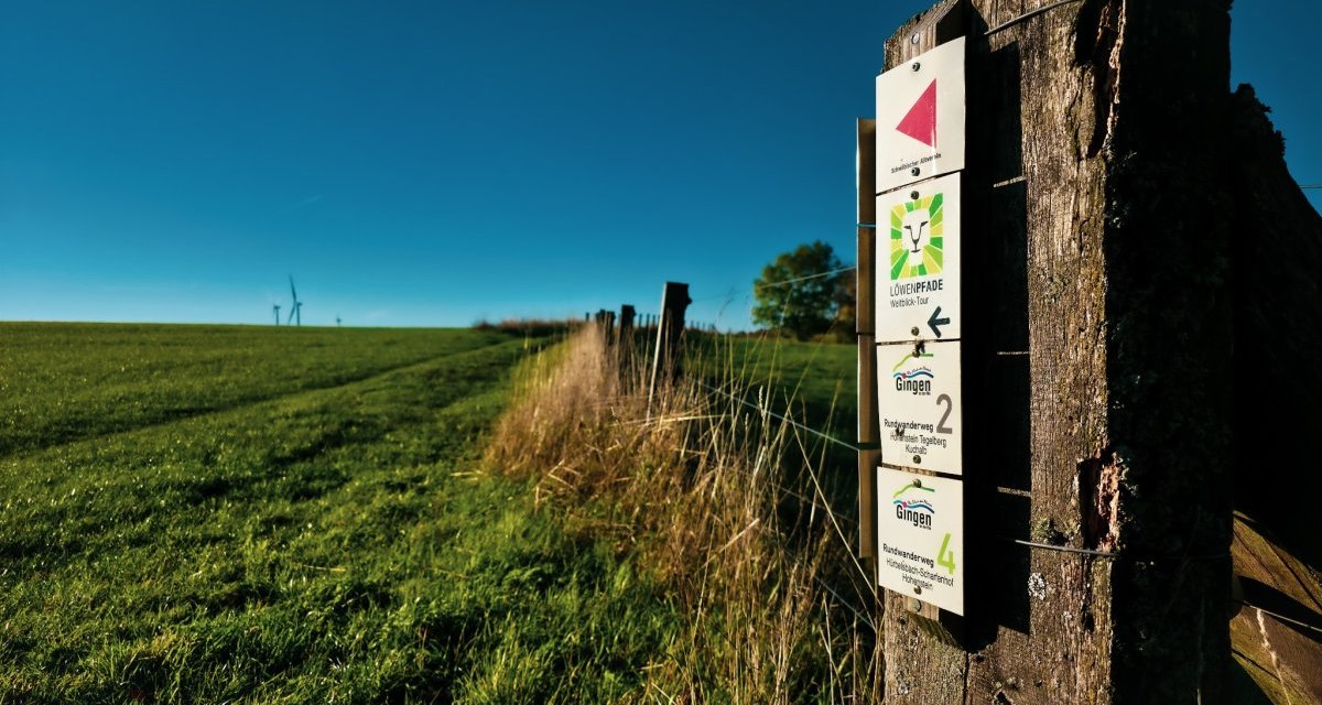 Ein Holzpfosten mit Wanderschildern steht auf einer grünen Wiese. Der Himmel ist klar und blau, im Hintergrund sind Windräder sichtbar., © Landkreis Göppingen Ein Holzpfosten mit Wanderschildern steht auf einer grünen Wiese. Der Himmel ist klar und blau, im Hintergrund sind Windräder sichtbar., © Landkreis Göppingen