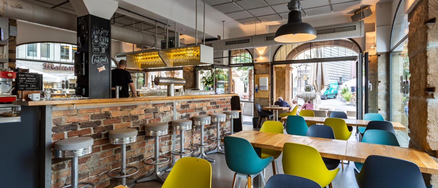 Modernes Café mit Bar aus Backstein, bunten Stühlen und großen Fenstern. Ein Mann steht hinter der Bar, draußen sind Passanten zu sehen., © deli, Stuttgart