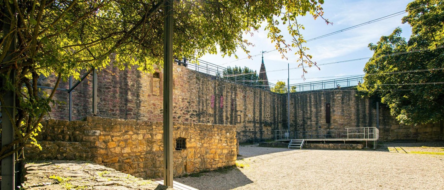 Steinmauern einer Burg in Marbach am Neckar, umgeben von Bäumen und Sonnenschein. Ein kleiner Platz mit Kiesboden ist im Vordergrund zu sehen., © Stuttgart-Marketing GmbH, Sarah Schmid