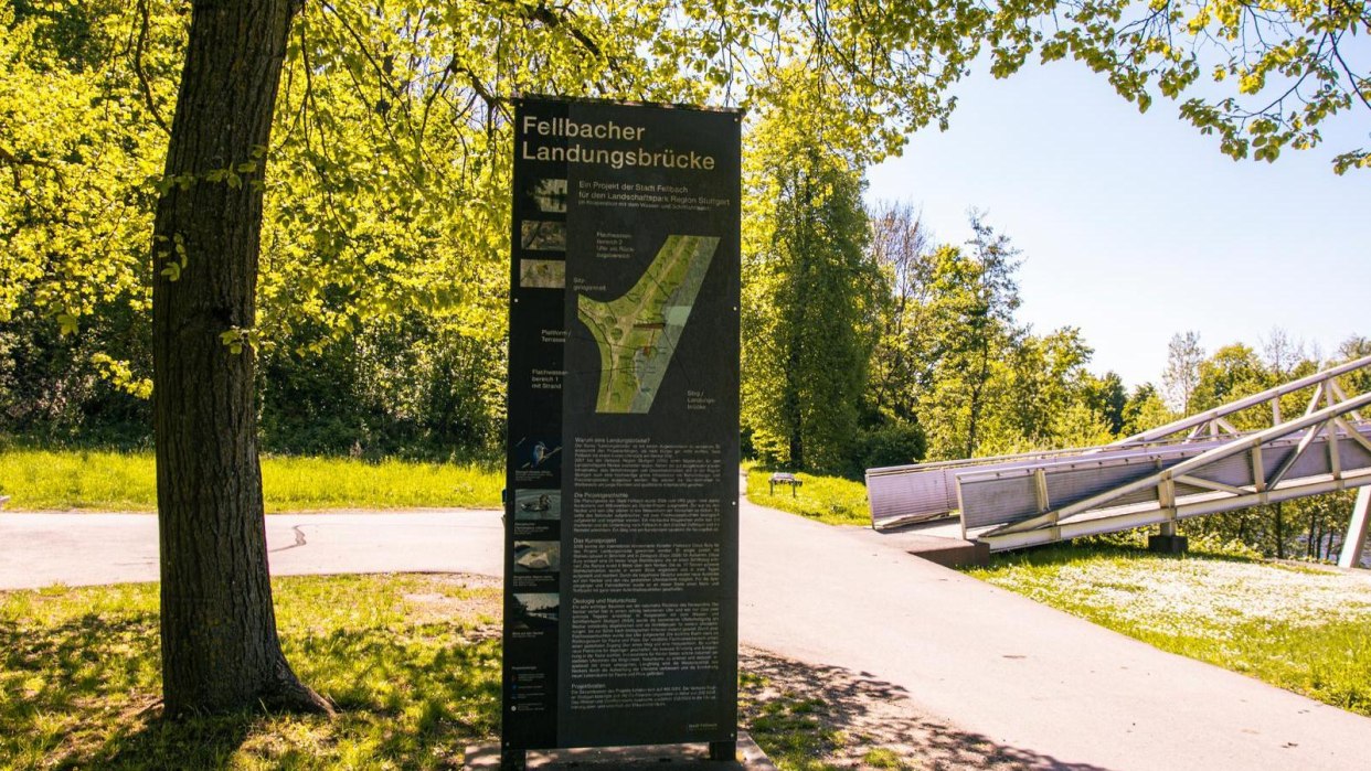 Sonniger Park mit Infotafel zur Fellbacher Landungsbrücke, umgeben von Bäumen und einem Gehweg. Eine Brücke ist im Hintergrund sichtbar., © Stuttgart-Marketing GmbH, Sarah Schmid Sonniger Park mit Infotafel zur Fellbacher Landungsbrücke, umgeben von Bäumen und einem Gehweg. Eine Brücke ist im Hintergrund sichtbar., © Stuttgart-Marketing GmbH, Sarah Schmid