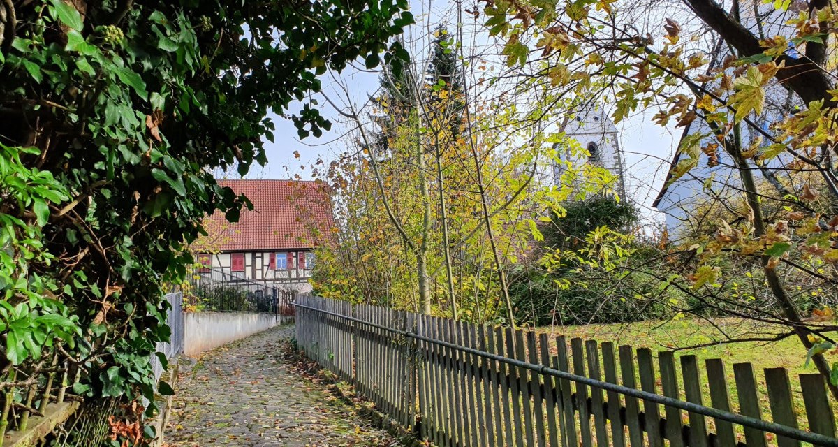 Ein gepflasterter Weg führt durch eine grüne Umgebung zu einem Fachwerkhaus und einem Kirchturm. Herbstliche Blätter und ein Holzzaun säumen den Weg., © Kulturamt Plochingen
