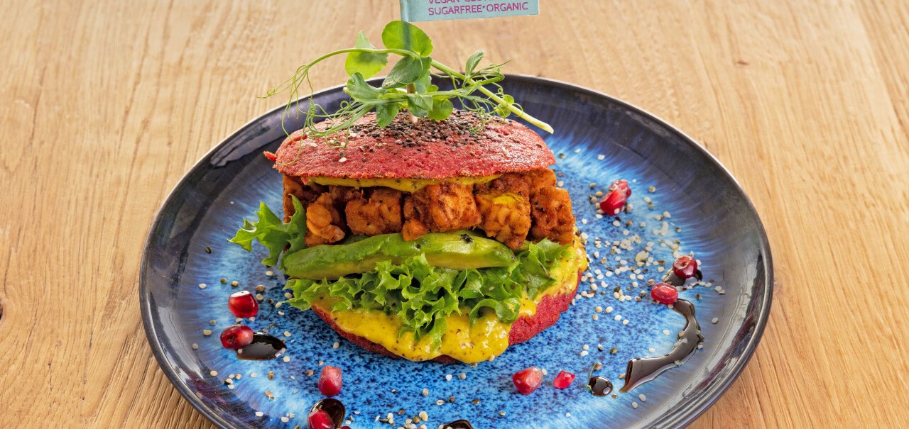 Ein veganer Burger mit Salat und Avocado auf einem blauen Teller, dekoriert mit Granatapfelkernen und Sesam. Ein F&auml;hnchen weist auf vegane, glutenfreie Zutaten hin., &copy; cEnergeticLife