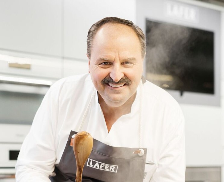 Ein Koch in wei&szlig;er Kleidung und Sch&uuml;rze mit der Aufschrift "Lafer" l&auml;chelt in die Kamera und h&auml;lt einen Holzl&ouml;ffel in einer modernen K&uuml;che., &copy; Stadthalle Leonberg