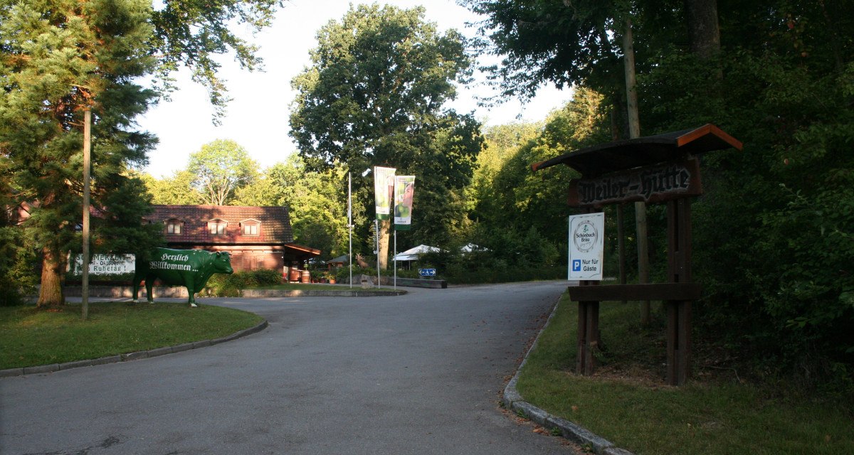 Eingang zur Weiler Hütte mit einem Schild und einer grünen Kuhfigur. Umgeben von Bäumen und einem Parkplatzschild für Gäste., © Natur.Nah. Schönbuch & Heckengäu Eingang zur Weiler Hütte mit einem Schild und einer grünen Kuhfigur. Umgeben von Bäumen und einem Parkplatzschild für Gäste., © Natur.Nah. Schönbuch & Heckengäu