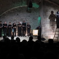 Ein Chor singt in einem Gewölbekeller. Ein Mann steht auf einer Leiter, während das Publikum zuschaut. Die Beleuchtung ist gedämpft., © Theater im Schlosskeller Ein Chor singt in einem Gewölbekeller. Ein Mann steht auf einer Leiter, während das Publikum zuschaut. Die Beleuchtung ist gedämpft., © Theater im Schlosskeller