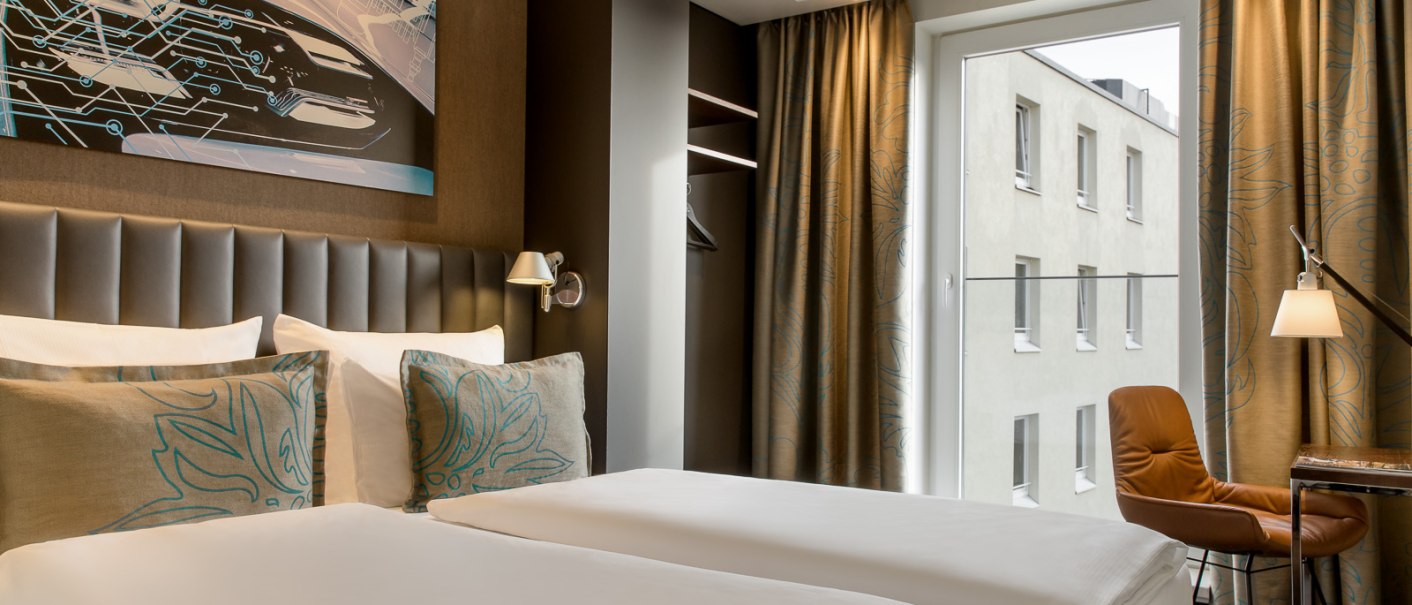 Modernes Hotelzimmer mit Doppelbett, Kunstwerk an der Wand, braunen Vorhängen und Fenster mit Blick auf ein Gebäude., © TOMAS