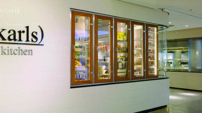 Eingang zu "karls kitchen" mit beleuchteten Glasvitrinen voller Lebensmittel und Blick in die moderne K&uuml;che., &copy; karls kitchen, Stuttgart