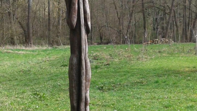 Eine abstrakte Holzskulptur steht auf einer grünen Wiese, umgeben von kahlen Bäumen und einem steinernen Hintergrund., © Natur.Nah. Schönbuch & Heckengäu Eine abstrakte Holzskulptur steht auf einer grünen Wiese, umgeben von kahlen Bäumen und einem steinernen Hintergrund., © Natur.Nah. Schönbuch & Heckengäu