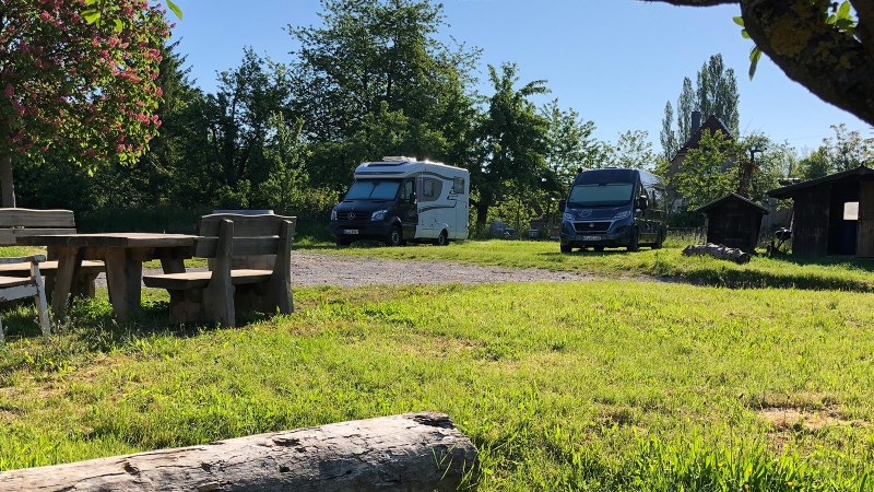 Wohnmobilstellplatz mit zwei Wohnmobilen, Holztisch und B&auml;nken unter einem bl&uuml;henden Baum. Im Hintergrund sind kleine Holzh&uuml;tten zu sehen., &copy; Bio Weingut Zimmer